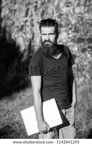 guy with long beard, hipster standing with laptop, computer, in tshirt on sunny, summer day on stone wall background. communication and new technology