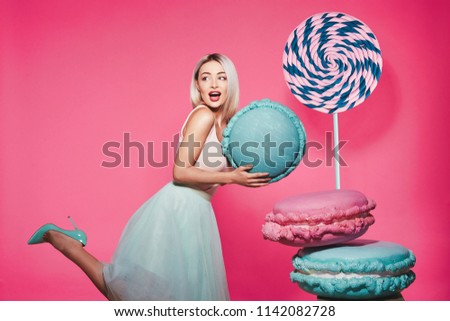 Cute young girl with blonde hair wearing top and skirt standing with huge sweet lollypop and macaroons at pink background, candy lover, Alice in Wonderland concept.
