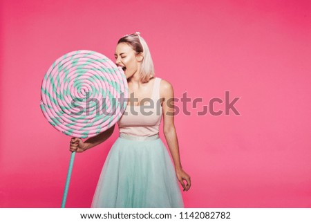 Pretty young girl with blonde hair wearing top and skirt standing with huge sweet lollypop and macaroons at pink background, candy lover, Alice in Wonderland concept.