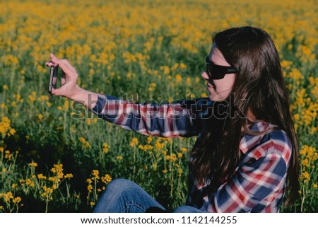 Woman makes a selfie on the phone sitting on the grass among the yellow flowers.