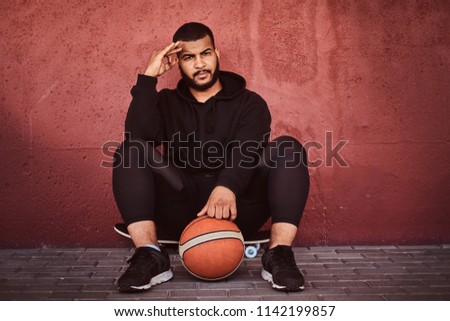 African-American bearded guy dressed in a black hoodie and sports shorts sitting on a skateboard with basketball and leaning on a wall.