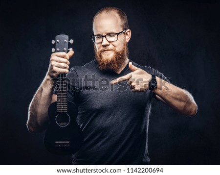 Redhead bearded male musician wearing glasses dressed in a gray t-shirt holds ukulele. Isolated on a dark textured background.