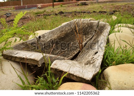 The old split wooden trough stands on round large smooth decorative stones. Bright green grass sucks through the stones.