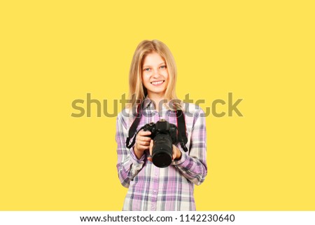 Young teenage girl holding digital photo camera with big lens & strap, taking pictures, smiling. Beautiful blond female photographer in checkered plaid shirt posing w/ dslr gear. Copy space background