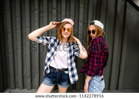 Two cheerful funny girl wearing checkered shirts posing against street wall at the street