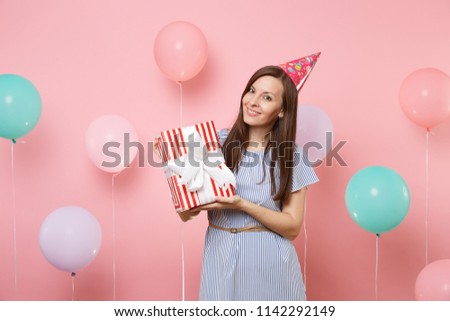 Portrait of fascinating young woman in birthday hat blue dress holding red box with gift present on pastel pink background with colorful air baloons. Birthday holiday party, people sincere emotions