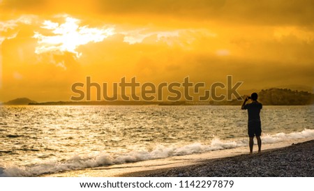 Man silhouette capturing the dramatic sunset on the beach. Beautiful blazing sunset landscape at blue sea and orange sky above it with awesome sun golden reflection on calm waves as a background. 