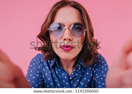 Funny young woman with bright makeup making selfie in studio. Indoor photo of good-looking female model in blue blouse taking picture of herself.