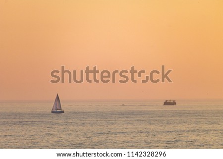 A distant sailboat sails along the sea during sunset, with the orange colored sky in the background.