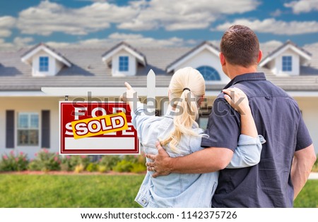 Young Adult Couple Facing and Pointing to Front of Sold Real Estate Sign and House.