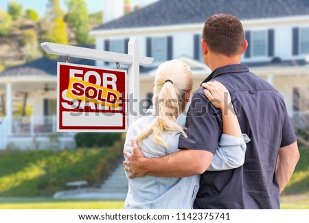 Young Adult Couple Facing Front of Sold Real Estate Sign and House.