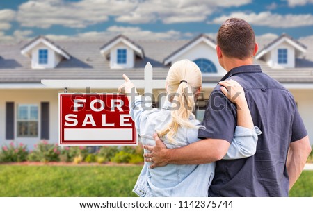 Young Adult Couple Facing and Pointing to Front of For Sale Real Estate Sign and House.