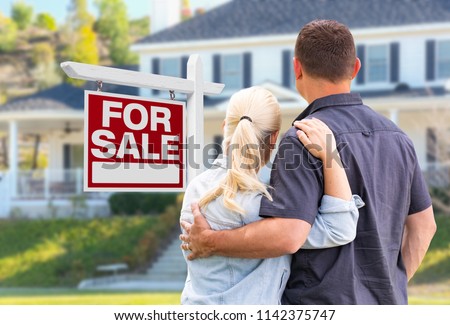 Young Adult Couple Facing Front of For Sale Real Estate Sign and House.