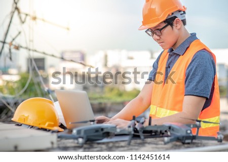 Young Asian engineer man working with drone laptop and smartphone at construction site. Using unmanned aerial vehicle (UAV) for land and building site survey in civil engineering project.