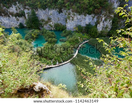 Plitvice Lakes National Park - UNESCO World Heritage Centre. Aerial view, colorful spring panorama of lit by sun green forest and long wooden bridge with tourists over blue lake with blue-green water.