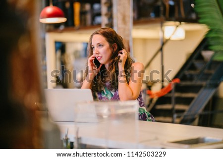 A young, attractive brunette woman sits at a counter in a cafe and is talking on her smartphone during the day. She is taking a break from work (her laptop is nearby) to have a conversation.