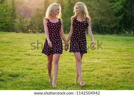 two sisters twins beautiful curly blonde happy young toothy smile women in stylish dress walking in the summer park sunset rays field background