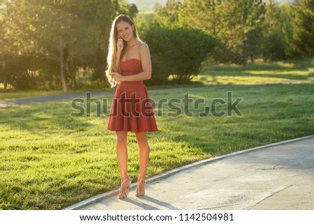 model woman in red dress on high stylish heels holds phone and talking in hand rays of sun background