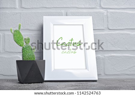
Small cactus in black concrete flower pot and mock-up of white frame with copy space for poster on gray brick wall background