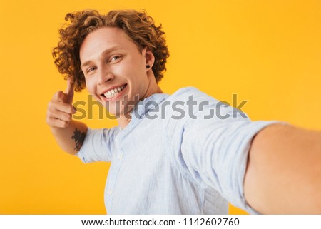 Photo of young beautiful man 20s taking selfie photo and pointing finger at camera isolated over yellow background