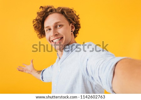 Portrait of astonished happy guy 20s taking selfie photo and holding copyspace on palm isolated over yellow background
