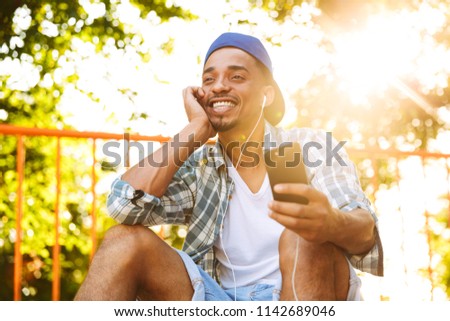 Cheerful young african man in earphones listening to music with mobile phone on a ramp at the skate park