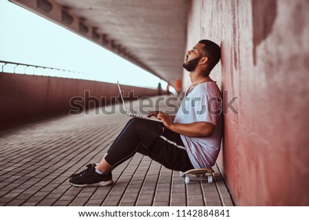 Pensive African-American bearded guy dressed in a white shirt and sports shorts holds on a laptop while sitting on a skateboard under a bridge, leaning on wall.