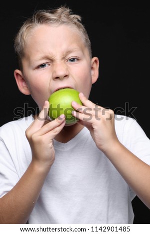 little blond boy bites into a green apple, looking at camera