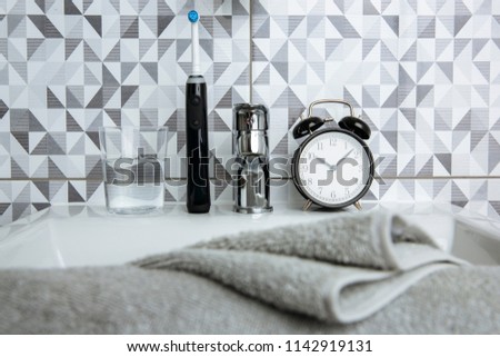 A black electric toothbrush, a clock and a glass of water standing on the bathroom sink