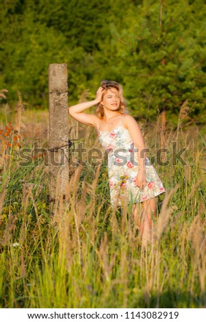 Beautiful young woman standing in a field near a concrete pillar, green grass and flowers. Outdoors Enjoy nature. Healthy smiling girl standing in tall grass