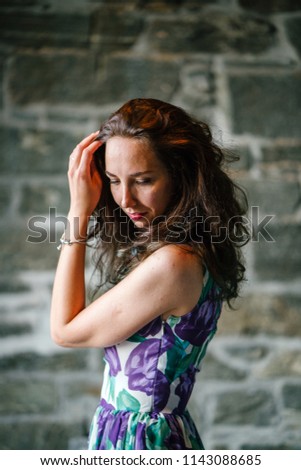 Portrait of a young, attractive Caucasian woman of European descent posing against a stone wall. She is tall, slim, brunette and is wearing a floral dress. 