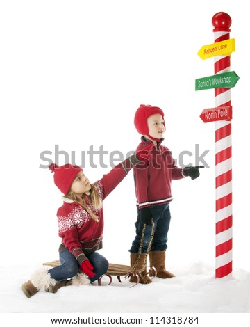 Two young children are on their way to the North Pole.  The sister points the way on one of Santa's directional signs.  On a white background with plenty of space for your text.