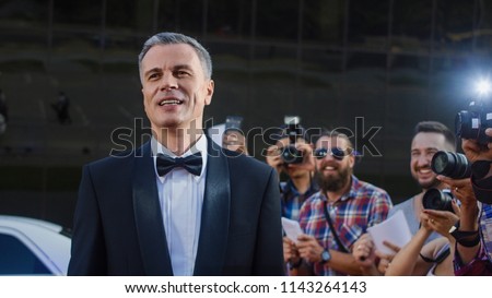 Handsome adult man in tuxedo standing on red carpet of luxury event at photographers and fans