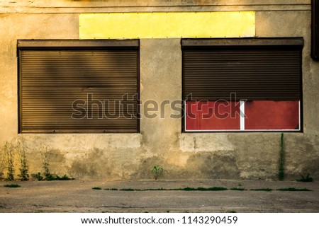 The building has two windows with rolling shutters and an empty signboard. One closed, the second half open