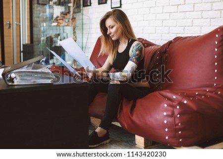 Tattooed girl sitting on sofa thoughtfully watching new tattoo sketches spending time in modern tattoo studio