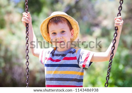 A cute Caucasian kid in a striped T-shirt and a straw hat playing on a swing.