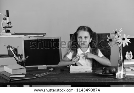 Schoolgirl with serious face writes in notebook in her classroom. Kid and school supplies on grey wall background. Girl sits at desk with colorful stationery, books and flowers. Back to school concept