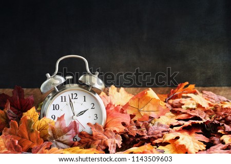 Alarm clock in colorful autumn leaves against a dark background with shallow depth of field. Daylight savings time concept.