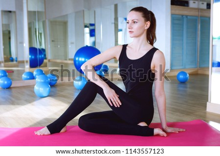 Woman sitting on pink ypga matt and doing exercises in fitness club gym, wearing black overall jumpsuit Royalty-Free Stock Photo #1143557123