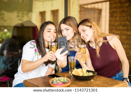Three young women sitting at restaurant with beers taking selfie with mobile phone
