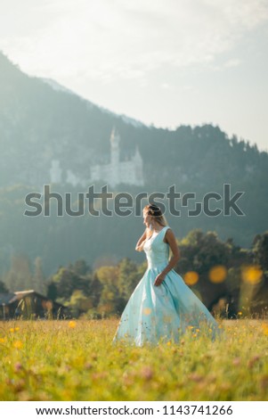 Pretty young woman in long blue dress dancing, smiling, having fun in green field with yellow flowers in Bavarian Alps, feeling happy and free. Copy space