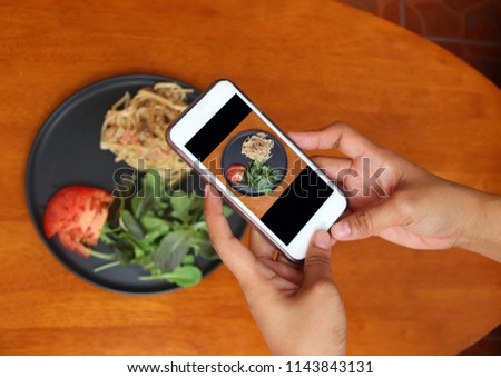 Hand with mobile phone take a photo of the The Spaghetti Carbonara with green vegetable in the black dish on the wooden table.
