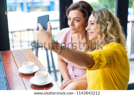 Smiling young women taking selfie photo and drinking coffee in cafe. Ladies sitting at table and posing. Women friendship and fun concept.