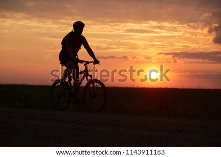Silhouette of a bicyclist traveling on a sunset background on the road to the field