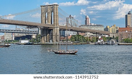 A sailing boat floating under the Brooklyn Bridge. View from New York City Lower Manhattan waterfront.