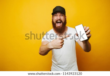 Amazed young bearded man in white t-shirt pointing at phone  Royalty-Free Stock Photo #1143923120