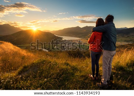 A young couple viewing sunset from the top of a mountain. The beautiful sun produces a warm orange glow to the entire image. Queenstown, New Zealand is a romantic getaway for lovers. Royalty-Free Stock Photo #1143934193