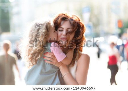 Attractive young woman with little charming girl are spending time together on sunny street. Girl kissing her mother and hugging and having fun. Happy family concept. 