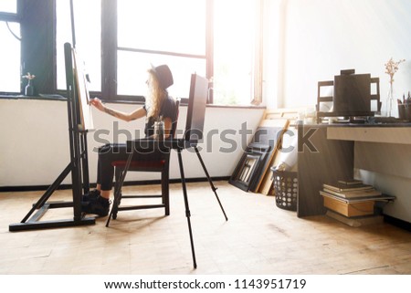 Tattoed amazing blonde curly girl in black hat drawing sketches behind the easel in studio near the sunny window