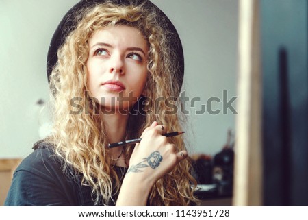 Portrait of long hair curly girl in handsome black hat sitting and dreaming with pencil behind the easel in studio